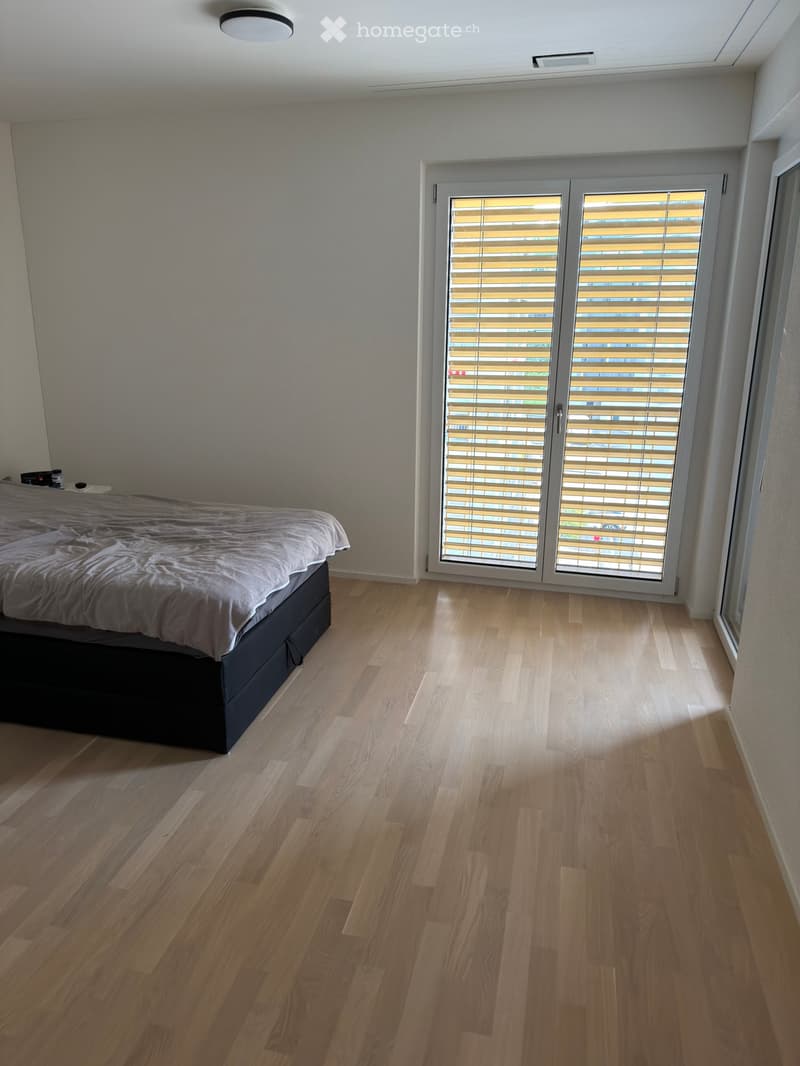 Modern bedroom with light wooden flooring, a neatly made bed with a grey duvet, and large sliding glass doors with blinds leading to a balcony.