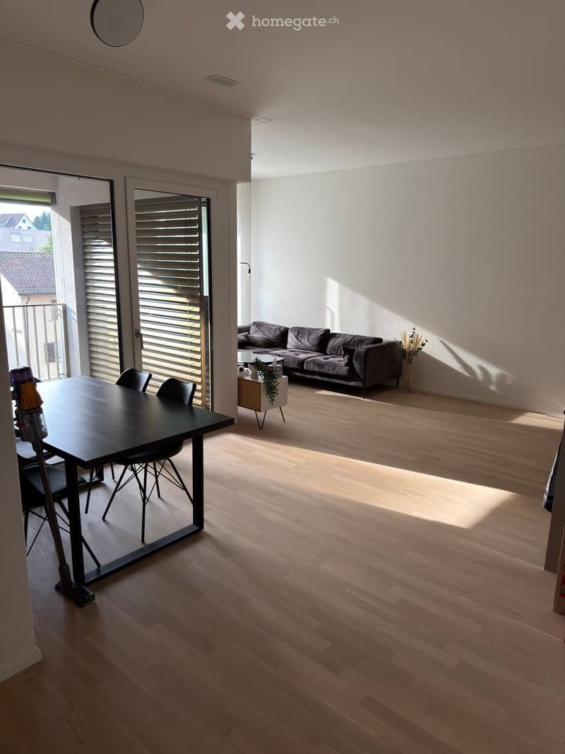 Open-plan living and dining area with light wooden flooring, a grey sofa, a black dining table with chairs, and a small side table with a plant and vase.