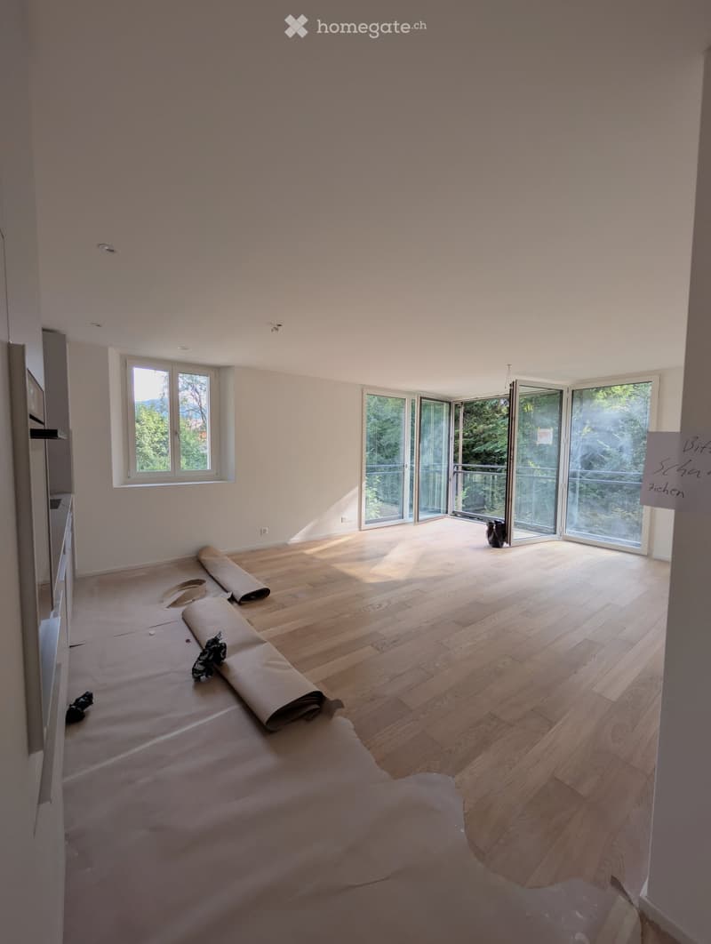 Empty room with wooden flooring, large windows, and sliding glass doors leading to a balcony. Some rolled-up materials and tools are visible on the floor.