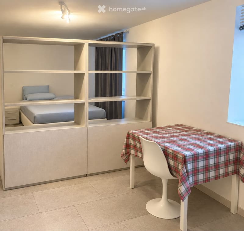 A compact, modern bedroom with built-in bunk beds, a small table with a plaid tablecloth, and a white chair. The room features light-colored walls, tiled flooring, and built-in shelving units.