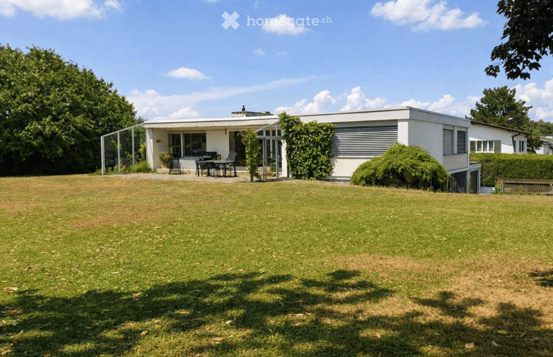 Single-story house with a flat roof, white exterior, and large windows. The house is surrounded by a well-maintained lawn and trees, with a small patio area featuring a table and chairs.
