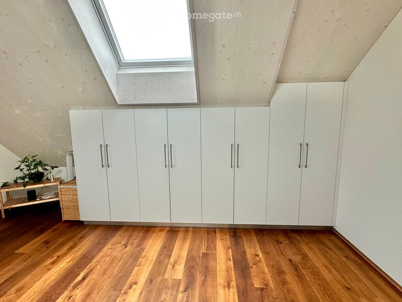 A modern, minimalist room with light-colored wooden flooring and white built-in cabinets. A small wooden table holds a potted plant and a wicker basket, adding a touch of greenery and texture to the space.