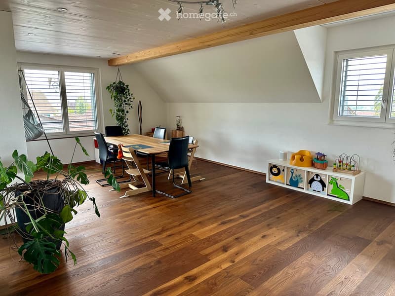 Bright, open room with wooden flooring, large windows, a dining table with chairs, a shelf with toys, and plants.
