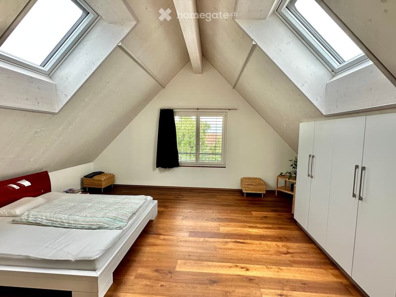 Bright attic bedroom with wooden floors, skylights, and large windows. Features a bed with a red headboard, a white wardrobe, two wicker stools, and a small table with a plant.