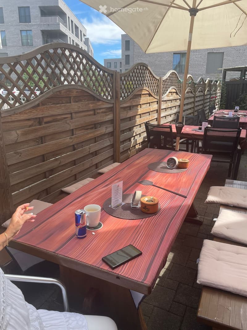 An outdoor seating area with wooden tables and chairs, a large umbrella providing shade, and a cup and can on the table.