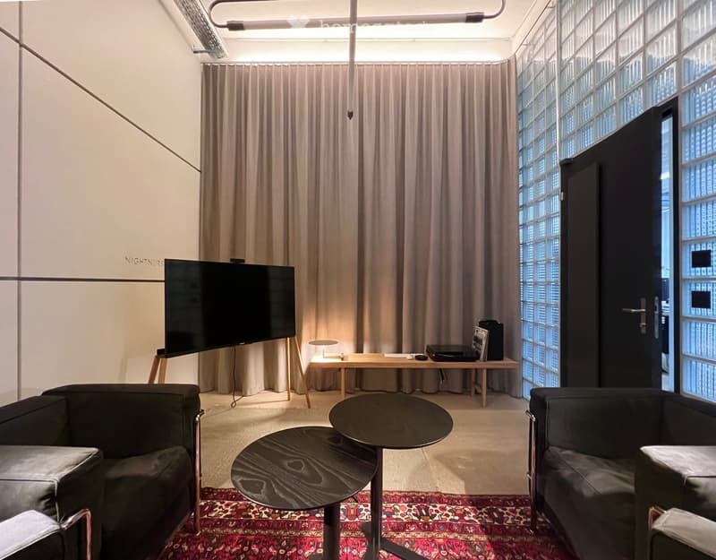 Modern living room with large windows, grey sofas, coffee tables, a screen, and a wooden desk with a lamp.