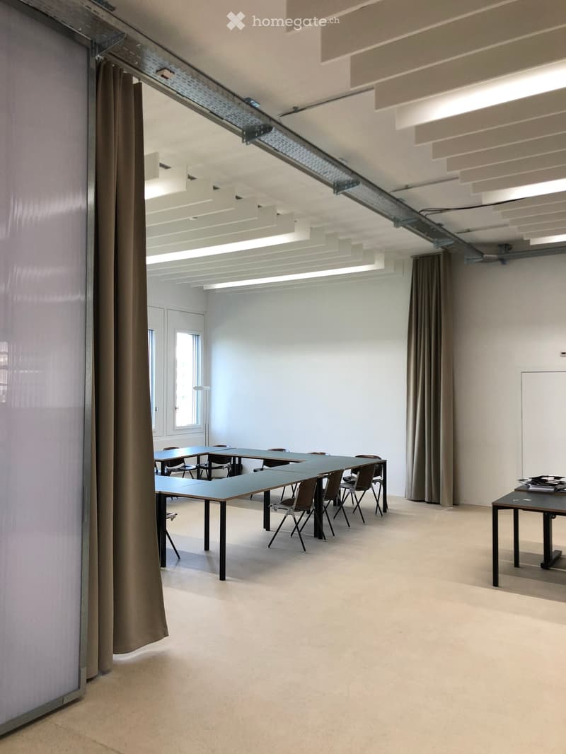Bright, open room with white walls and a high ceiling, featuring multiple tables and chairs arranged for meetings or collaborative work. Large windows and curtains provide natural light.