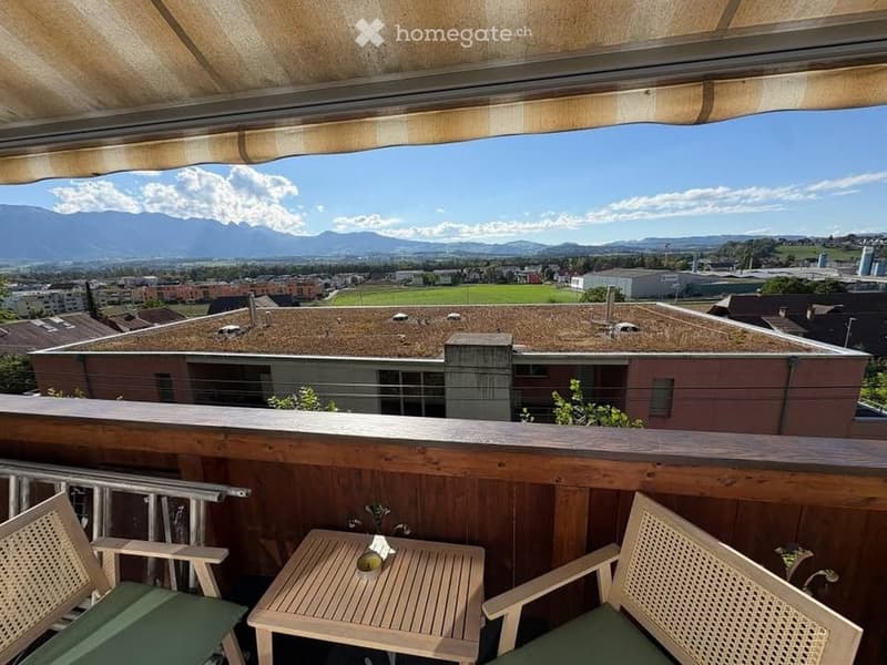 Balcony with wooden table and two chairs, overlooking a cityscape with mountains in the background.