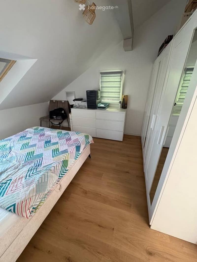Cozy attic bedroom with wooden flooring, slanted ceilings, and large windows. Features a single bed with a colorful geometric-patterned duvet, a white dresser, a small chair, and a mirror.