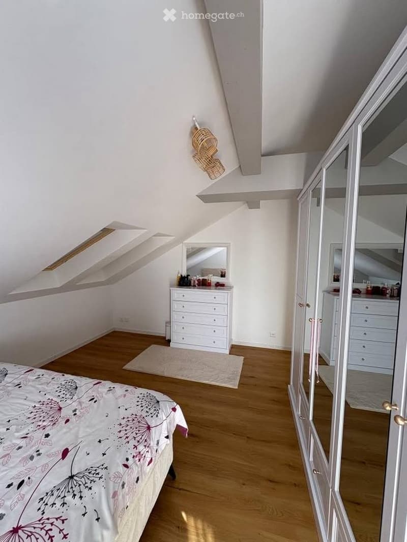 Bright attic bedroom with wooden flooring, slanted ceilings, and large windows. Features a bed with a floral duvet, a white dresser, a mirror, and a walk-in closet with a mirrored door.