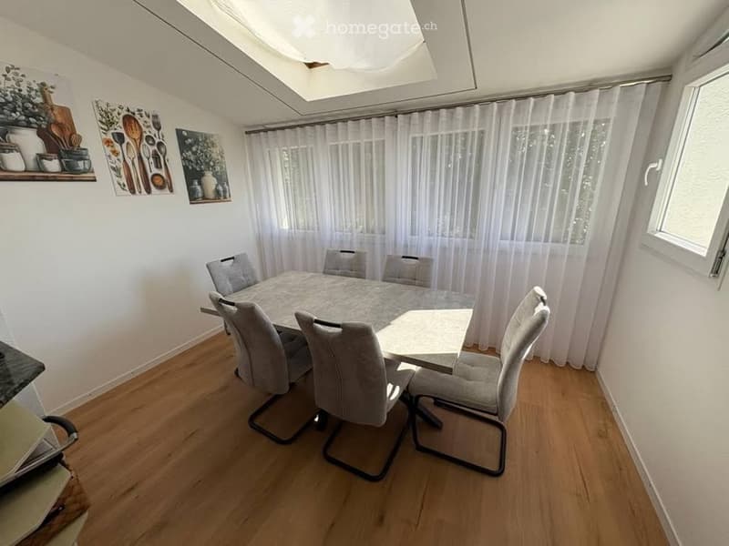 Dining area with a round table and four chairs, wooden flooring, white walls, and large windows with white curtains. Shelves with decorative items and artwork are visible on the wall.
