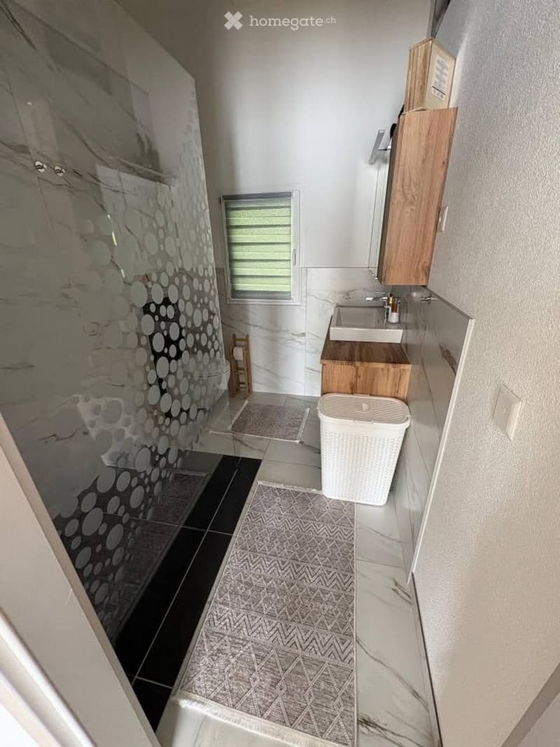 Modern bathroom with a wooden sink cabinet, mirror, and a laundry basket. The floor features a geometric patterned tile design, and there is a glass-enclosed shower area with a decorative tile backsplash.