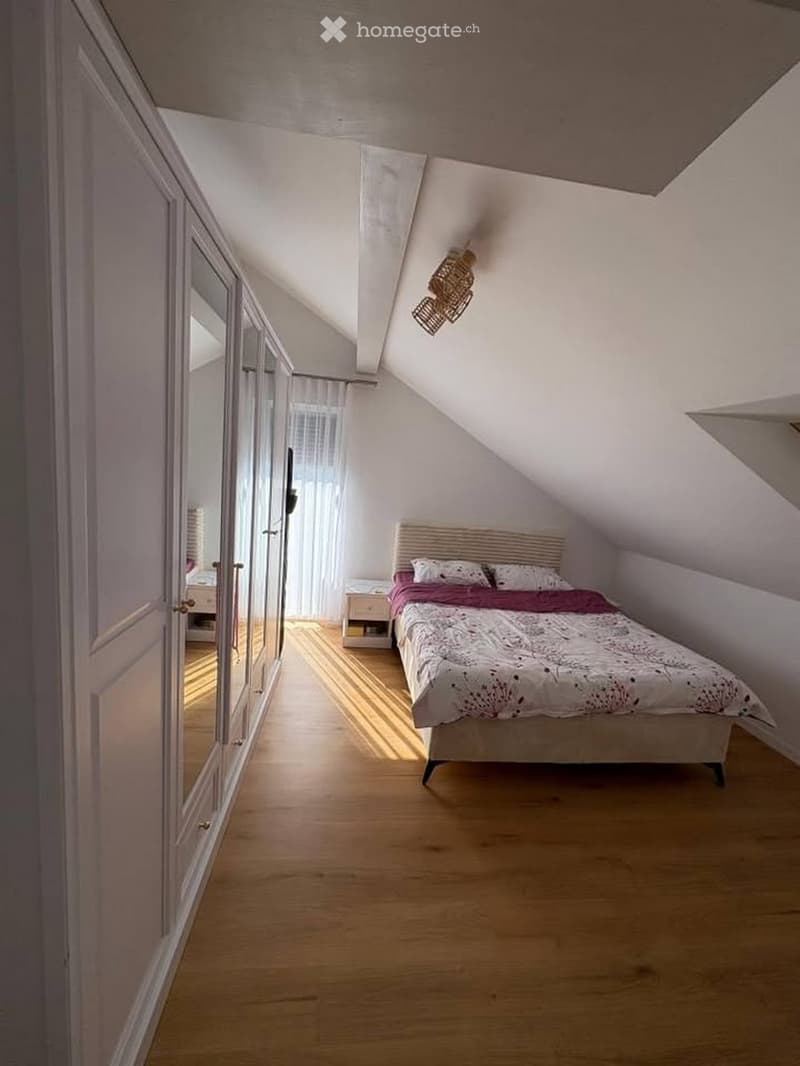 Cozy attic bedroom with wooden flooring, slanted ceilings, a bed with a decorative headboard, a nightstand, and a large mirrored wardrobe.