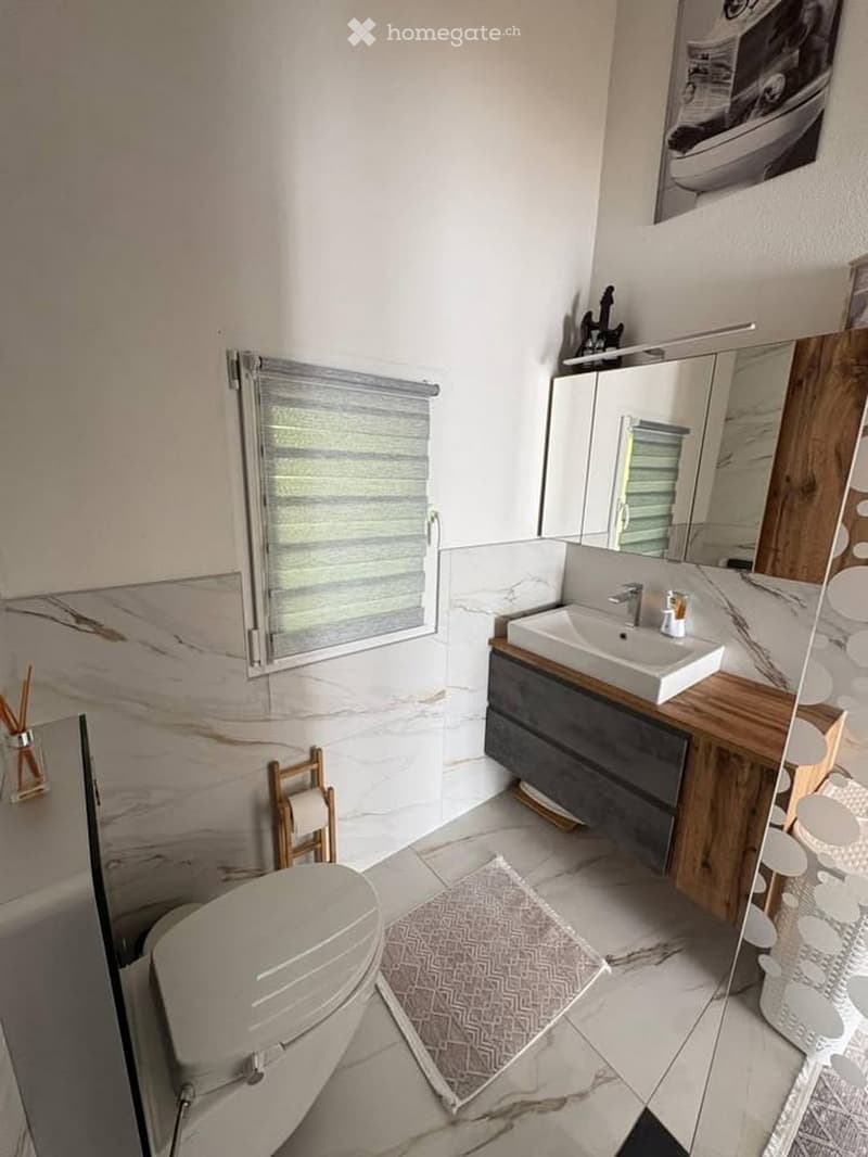 Modern bathroom with marble tiles, a white sink with wooden cabinet, a small wooden stool, a toilet, and a patterned rug on the floor.