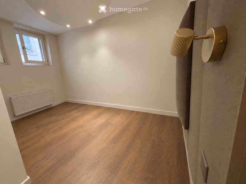 Empty room with wooden flooring, white walls, a window, a radiator, and a modern wall-mounted light fixture.