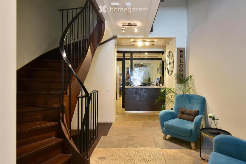 Spacious hallway with a wooden staircase, reception area, and seating area featuring a blue armchair and small table.