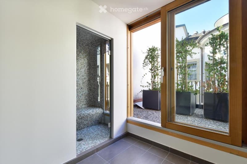 Balcony with large sliding glass doors, view of potted plants and neighboring buildings, tiled floor, and adjacent shower area visible through an open door.