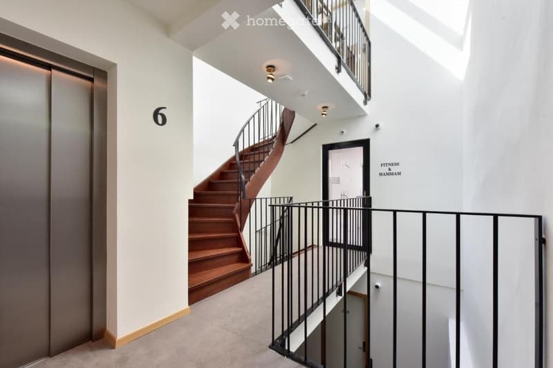 Modern hallway with a wooden staircase, black metal railing, elevator door marked with number 6, and a sign for 'Fitness Hammam'.