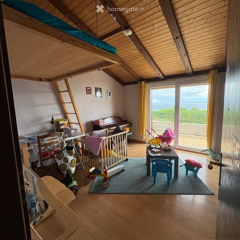Cozy attic room with wooden floors, slanted ceilings, and large windows offering a scenic view. Features a desk, chair, small table with toys, crib, piano, and a ladder leading to a lofted sleeping area.