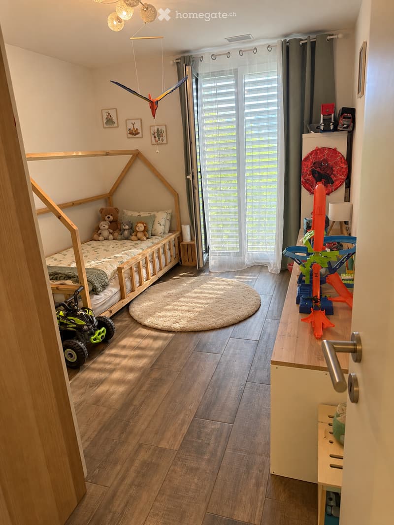 A well-lit children's bedroom with a wooden bed, plush toys, a round rug, and various toys including a toy car and a toy airplane. The room features wooden flooring, large windows with blinds, and a hanging toy airplane.
