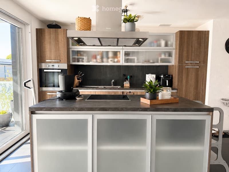 Modern kitchen with wooden cabinets, stainless steel appliances, and a dark countertop. Features include a built-in oven, refrigerator, coffee maker, sink, and various kitchen accessories.