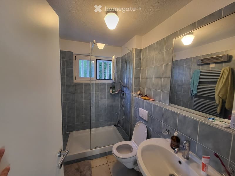 Modern bathroom with grey tiled walls, a bathtub, toilet, sink, mirror, and a glass-enclosed shower area. The room features a window with blinds and a towel hanging on a radiator.