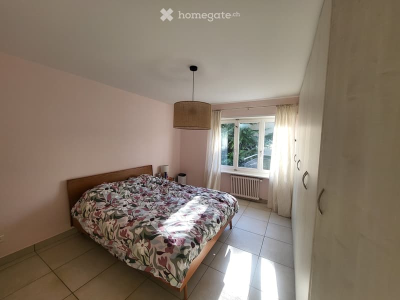 Bedroom with a double bed, nightstand, lamp, and window with curtains. Light pink walls and tiled floor.