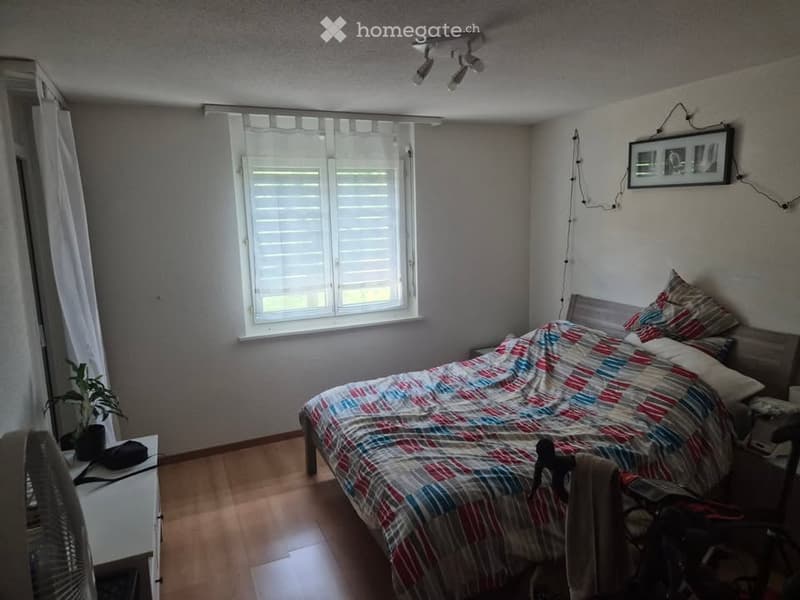 Bedroom with wooden flooring, a bed with a colorful quilt, a nightstand, a fan, a small plant, and a picture frame on the wall.