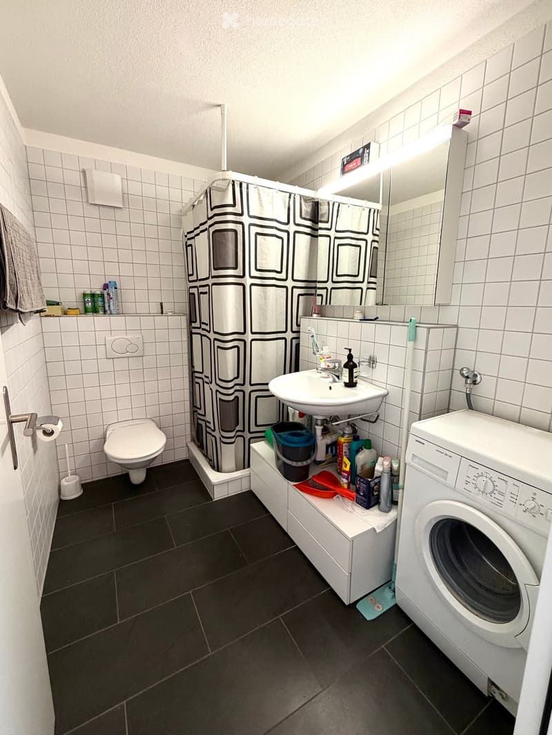 Modern bathroom with white tiles, a toilet, a sink with a mirror, a washing machine, and a shower area with a black and white patterned curtain.