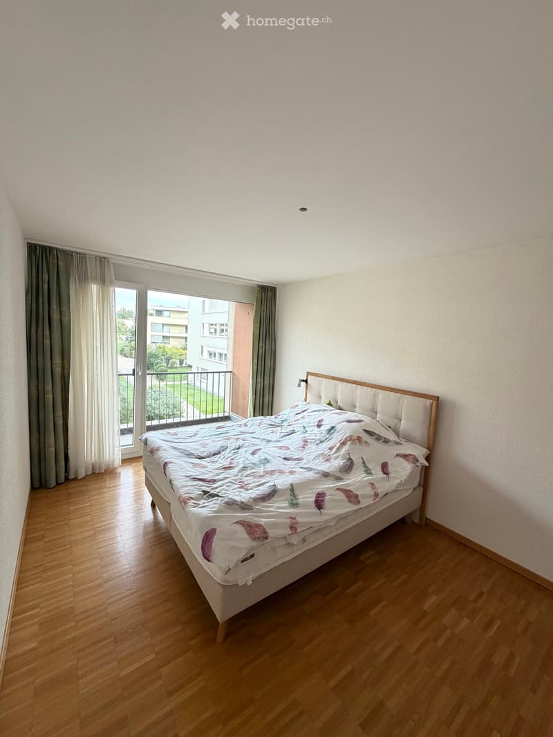 Bedroom with wooden flooring, a bed with a feather-patterned duvet, and a balcony with a view of greenery.