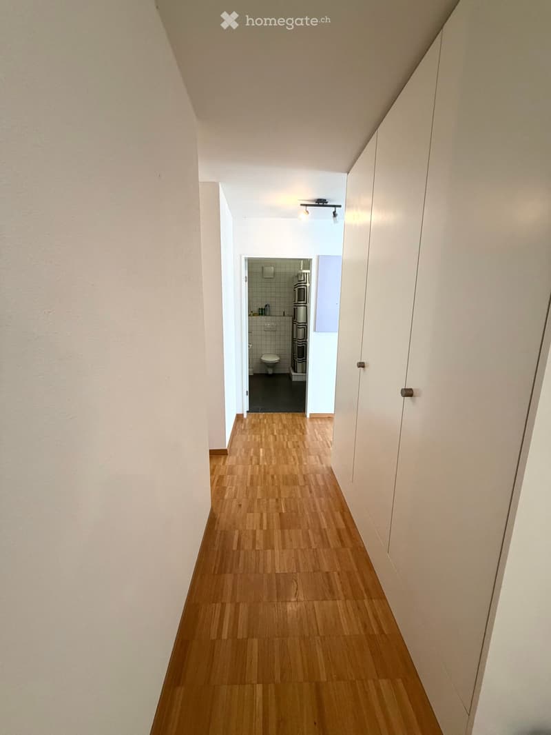 A narrow hallway with wooden flooring and white walls, leading to a small bathroom with a toilet, shower, and tiled walls.