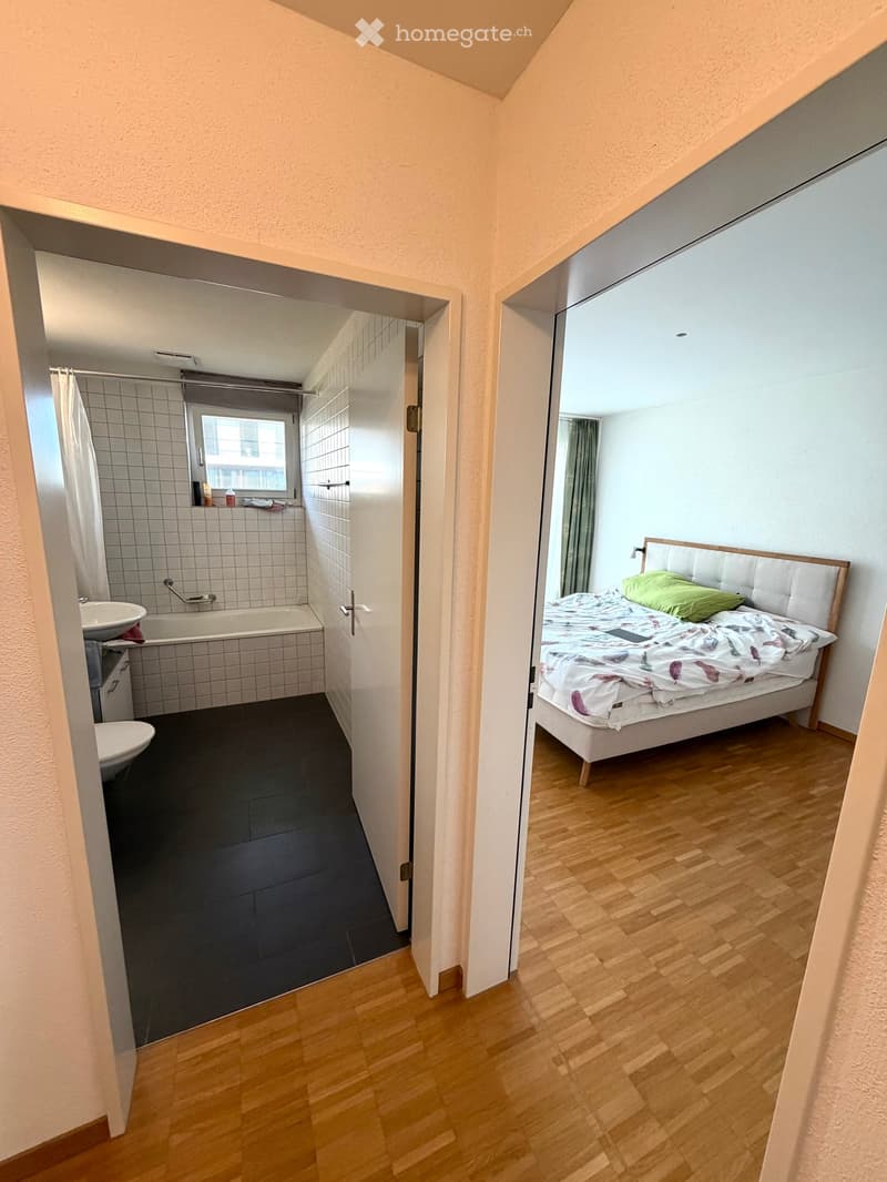A modern bedroom with a wooden floor, a white headboard bed, and a green pillow. An adjacent bathroom with a bathtub, sink, and toilet is visible through an open doorway.