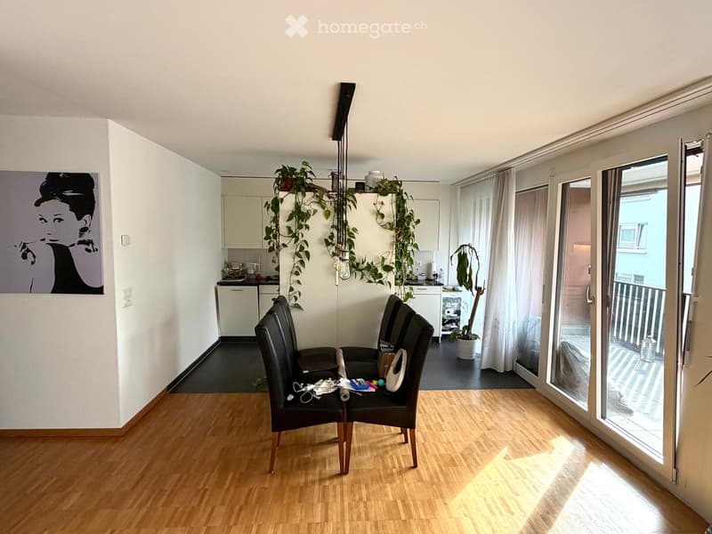 A modern living room with wooden flooring, two black chairs, a small table, kitchen area with cabinets, and a potted plant on the windowsill. Large windows and sliding doors lead to a balcony.