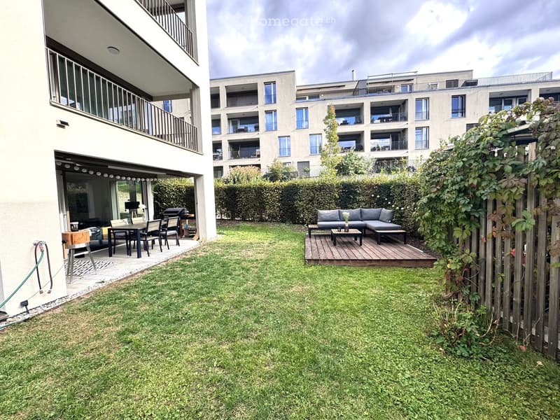 Well-maintained garden with a wooden deck, seating area, and a modern apartment building in the background.