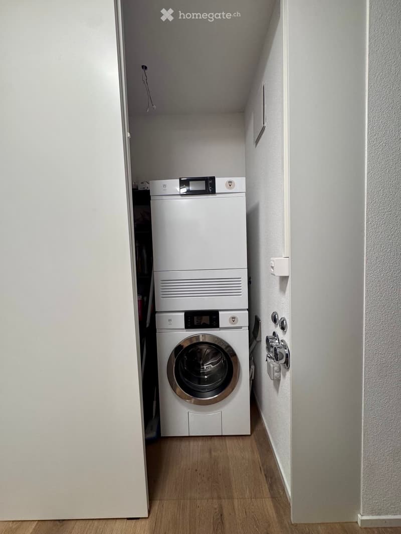 A small, narrow bathroom with a white washing machine and dryer stacked vertically, wooden flooring, and white walls.