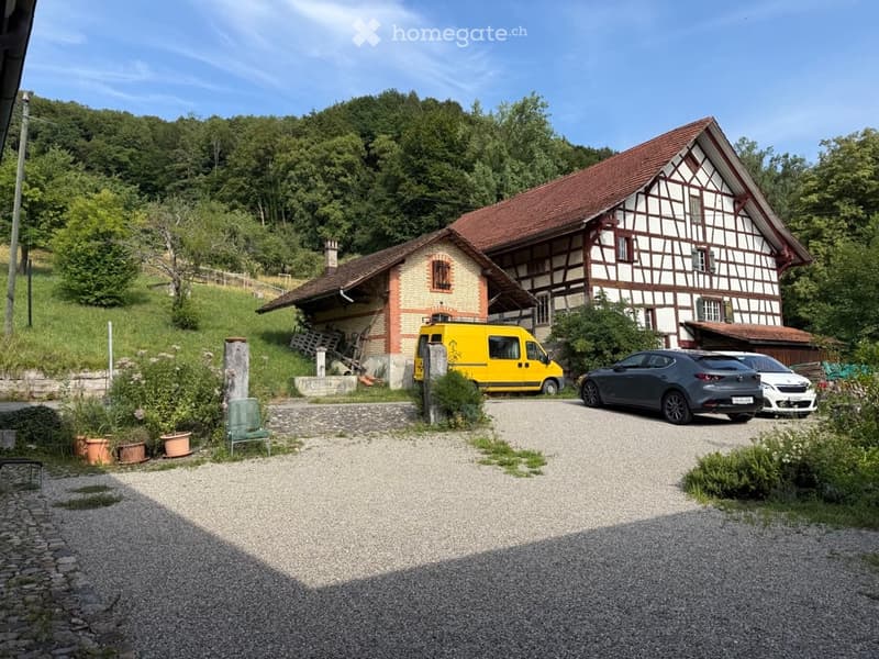 Traditional half-timbered house with red-tiled roof, surrounded by greenery, with a gravel driveway and parked vehicles.