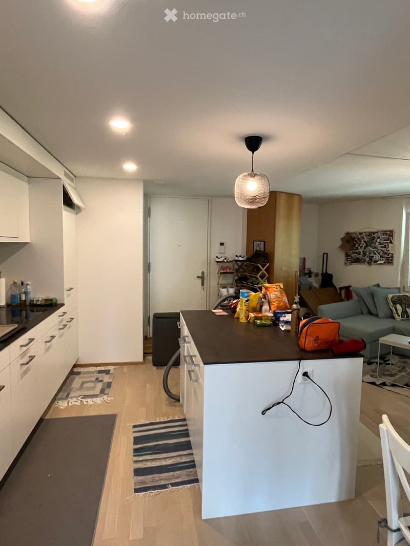 Modern kitchen and living area with white cabinets, dark kitchen island, pendant light, sofa, and various items on the counter.