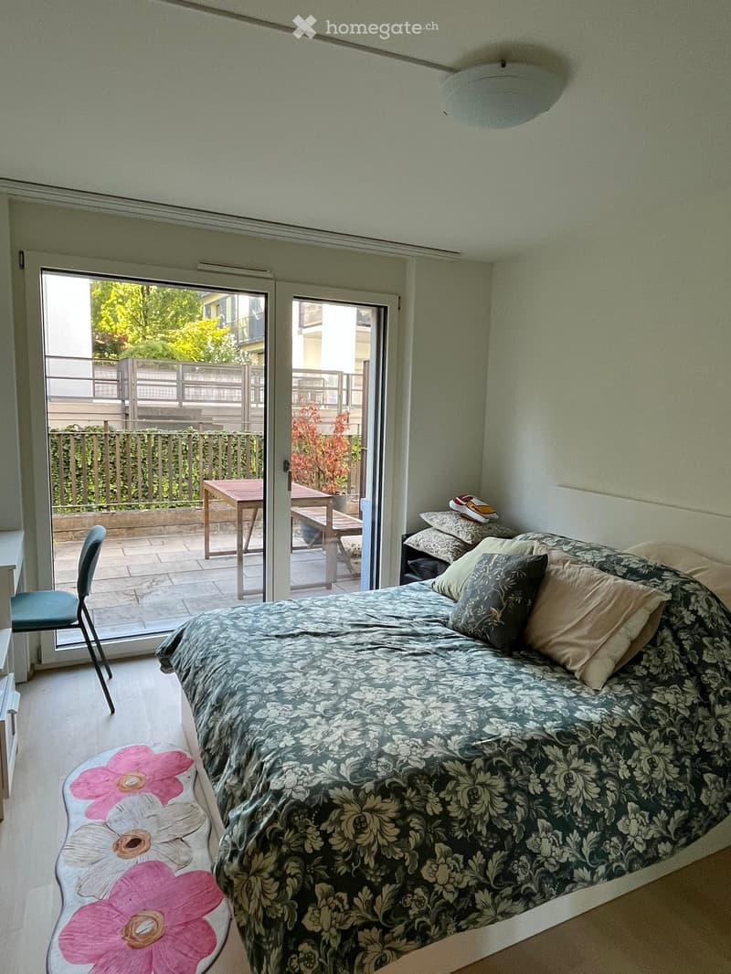 Bedroom with a large bed, patterned bedspread, pillows, a chair, a small table, and a round stool with a floral design near the bed.