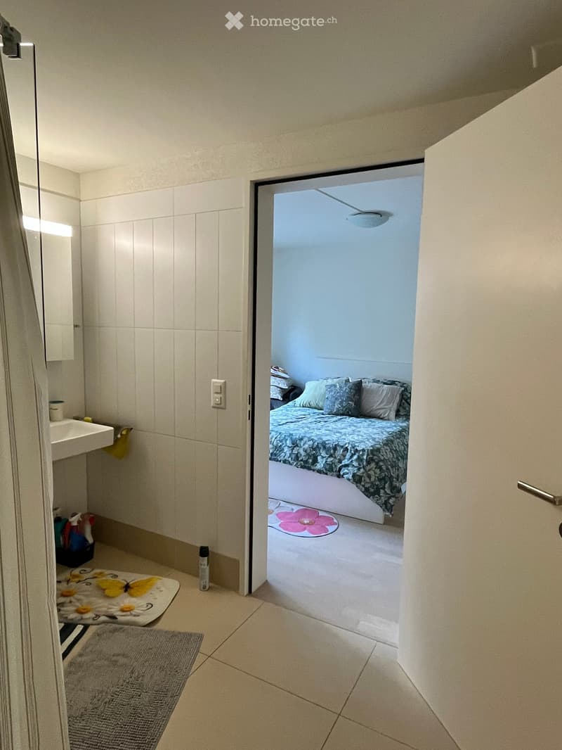 A small bathroom with a sink, mirror, and a rug, leading to a bedroom with a bed, pillows, and a blanket.