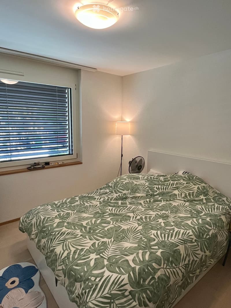 Bedroom with a single bed covered in a green leaf-patterned duvet, a small stool with a blue flower design, a wall-mounted fan, and a window with blinds.