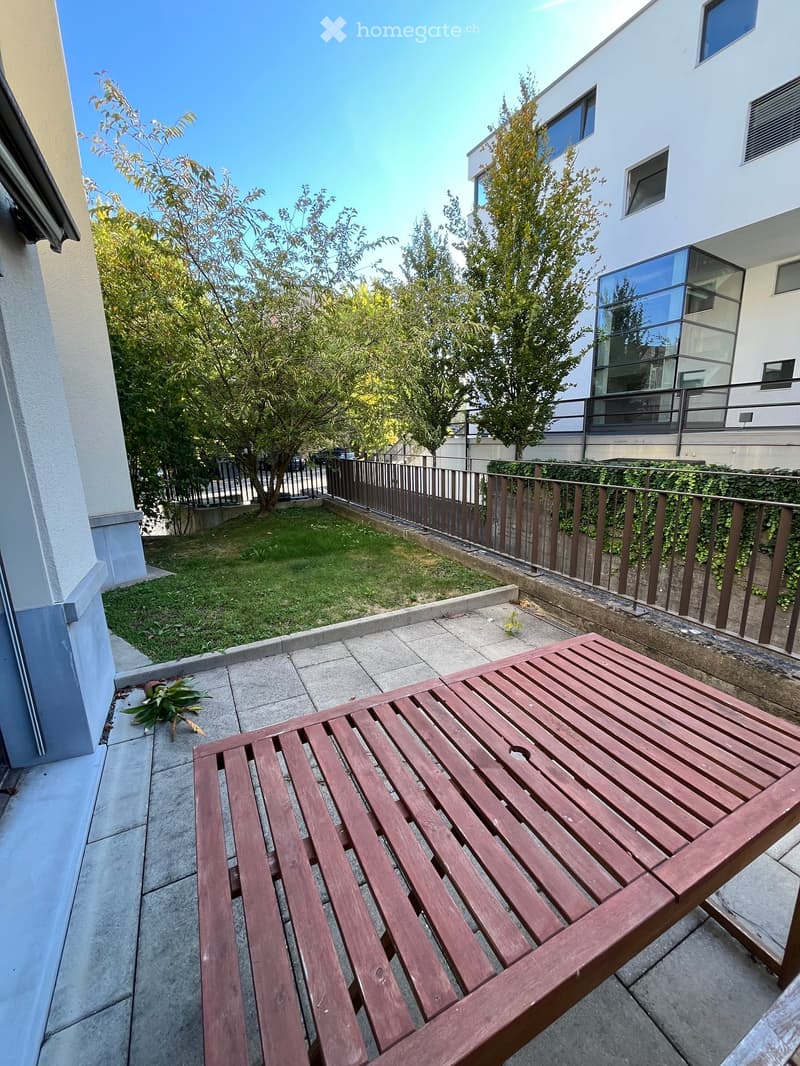 Balcony with a wooden table, small potted plant, and a view of a fenced garden area with trees and a modern building.