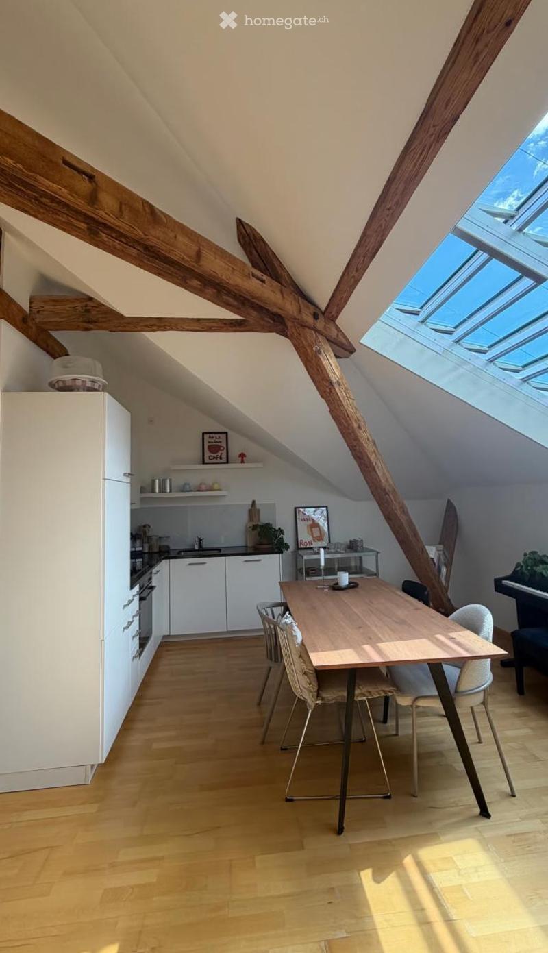 Bright attic space with wooden beams, skylight windows, and a combined kitchen and dining area. Features a wooden dining table with chairs, white kitchen cabinets, and a piano in the corner.