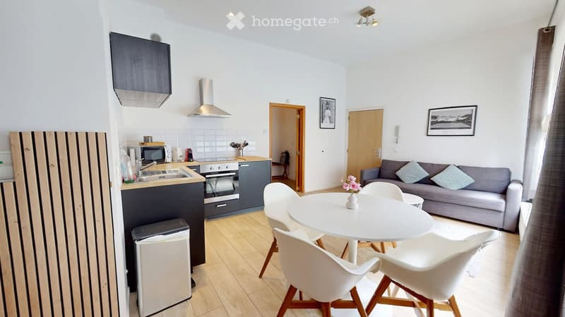 A modern, open-plan living and dining area with a grey sofa, white dining table, and four white chairs. The kitchen features dark cabinets, a microwave, oven, and sink. The space includes a trash bin and picture frames on the walls.
