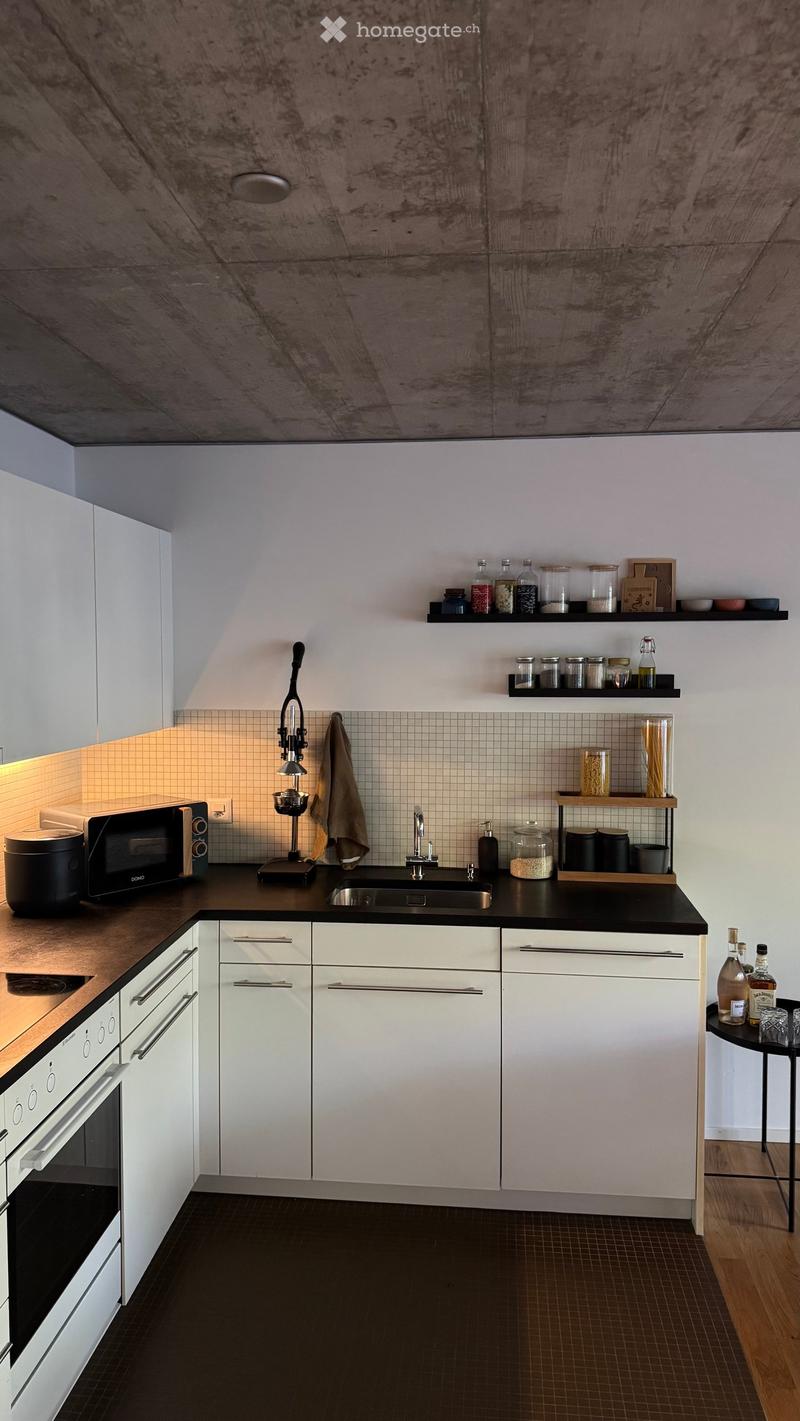 Modern kitchen with white cabinets, black countertops, stainless steel sink, microwave, oven, and coffee machine. Shelves hold jars and bottles, and a small round table with bottles and glasses is visible.