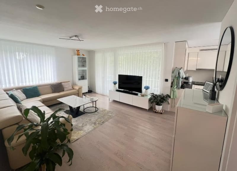 Bright living room with a beige sectional sofa, coffee table, TV stand, and a small cabinet. The room features large windows with white curtains and a wooden floor.