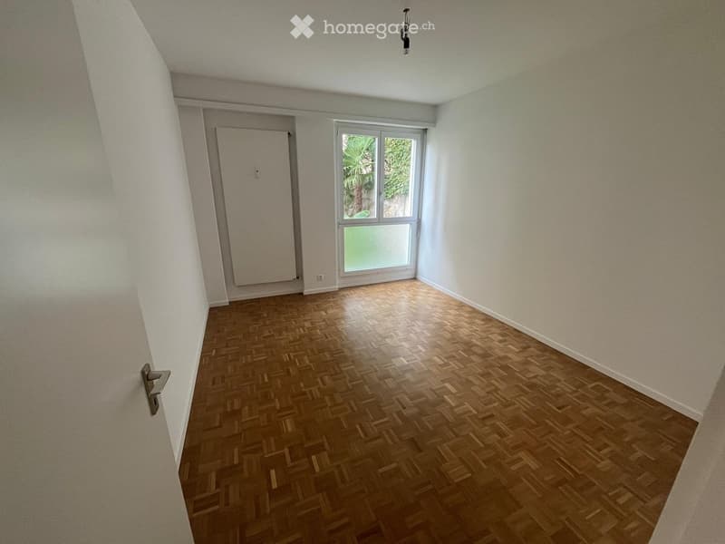 Empty room with wooden parquet flooring, white walls, and a large window with a view of greenery outside.