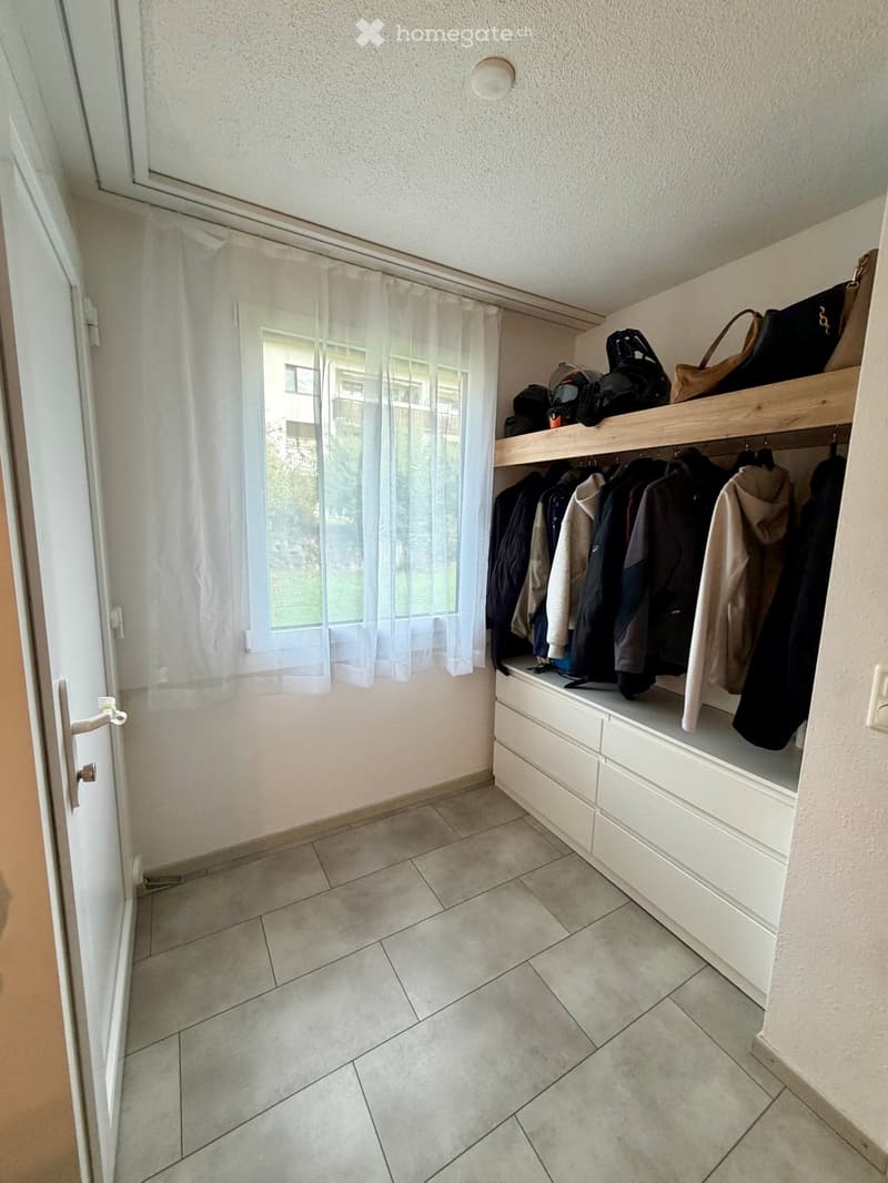 A well-lit hallway with tiled flooring, white built-in storage unit with drawers and hanging space for coats, and a window with sheer curtains allowing natural light.