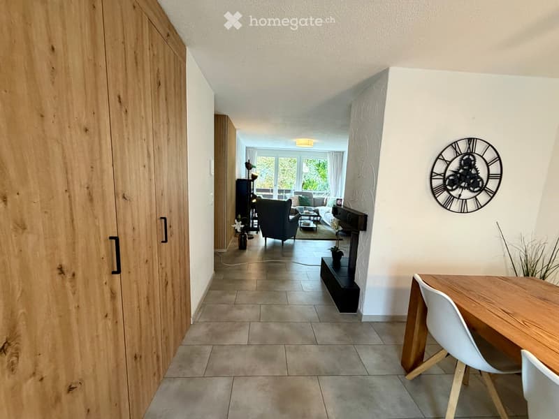 Modern hallway leading to a living area with a sofa, armchair, coffee table, and a dining area with a table and chairs. A large wall clock is mounted on the wall.