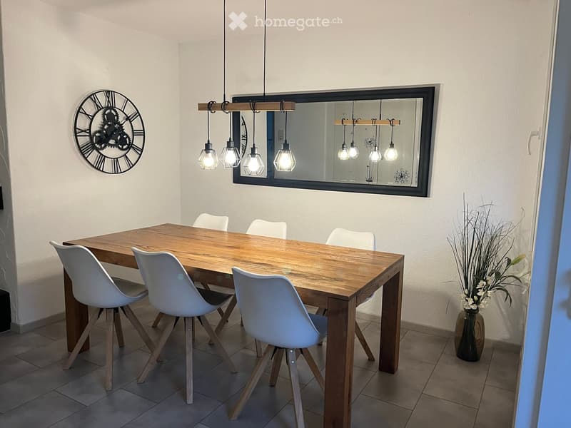Dining room with a wooden dining table, white chairs, a vase with flowers, a large mirror, and a wall clock.
