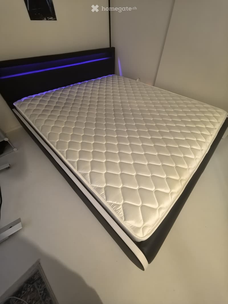 A modern bedroom with a white quilted mattress on a black bed frame, illuminated by blue LED lighting along the headboard.